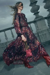 Model in a floral sheer maxi dress and red boots holding a pink leather handbag against a stone balcony