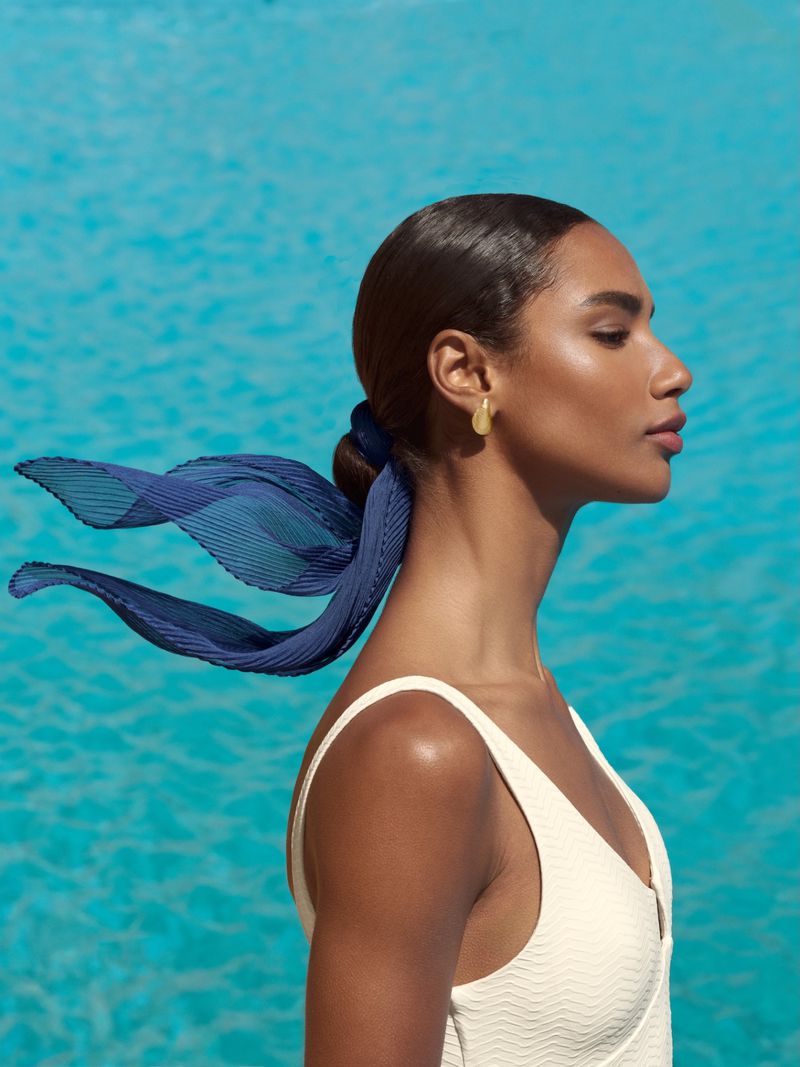 Model in white swimwear with blue hair ribbon and gold earrings against sparkling blue water for Sofia Godinho S/S 26