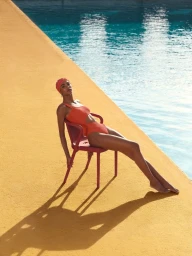 Model seated on a red chair poolside wearing an orange swimsuit and matching swim cap