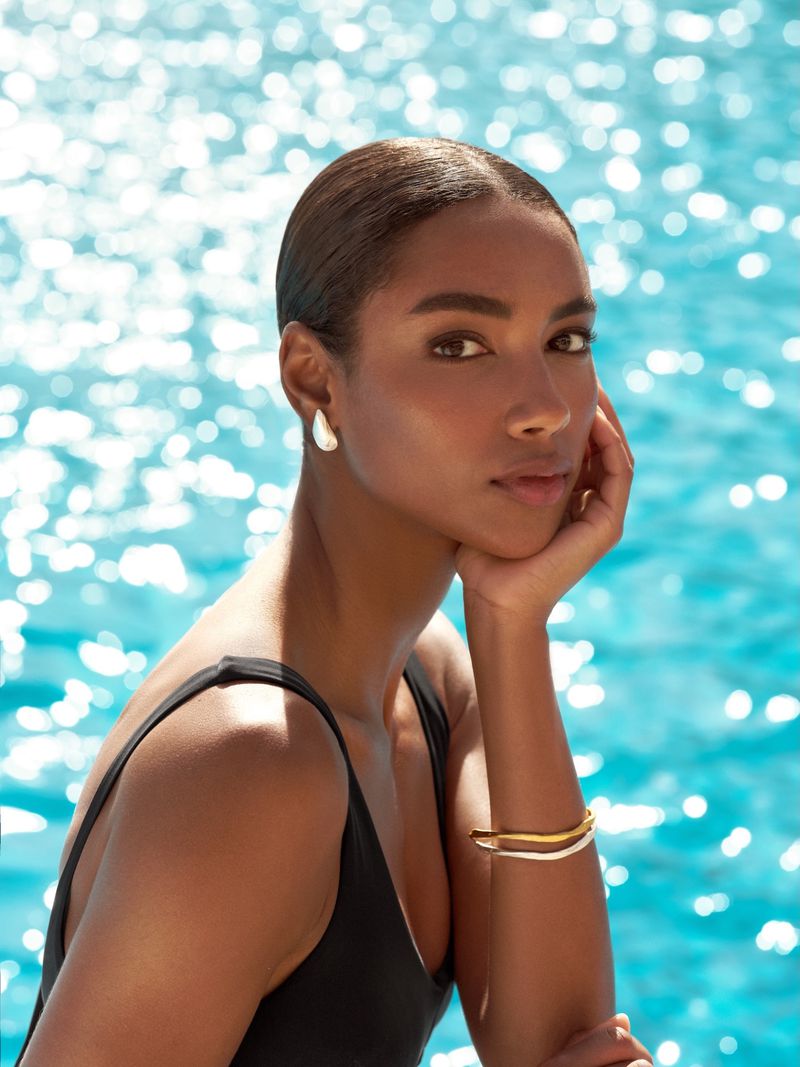 Model wearing a black swimsuit and gold jewellery posing by a glittering blue pool water background