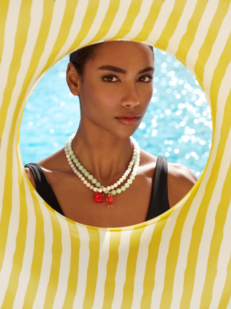 Model wearing layered beaded necklaces and a black swimsuit framed inside a yellow and white striped pool float
