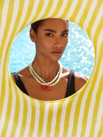 Model wearing layered beaded necklaces and a black swimsuit framed inside a yellow and white striped pool float