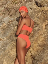 Model in an orange swimsuit and matching swim cap posing against a rugged rocky cliff face.