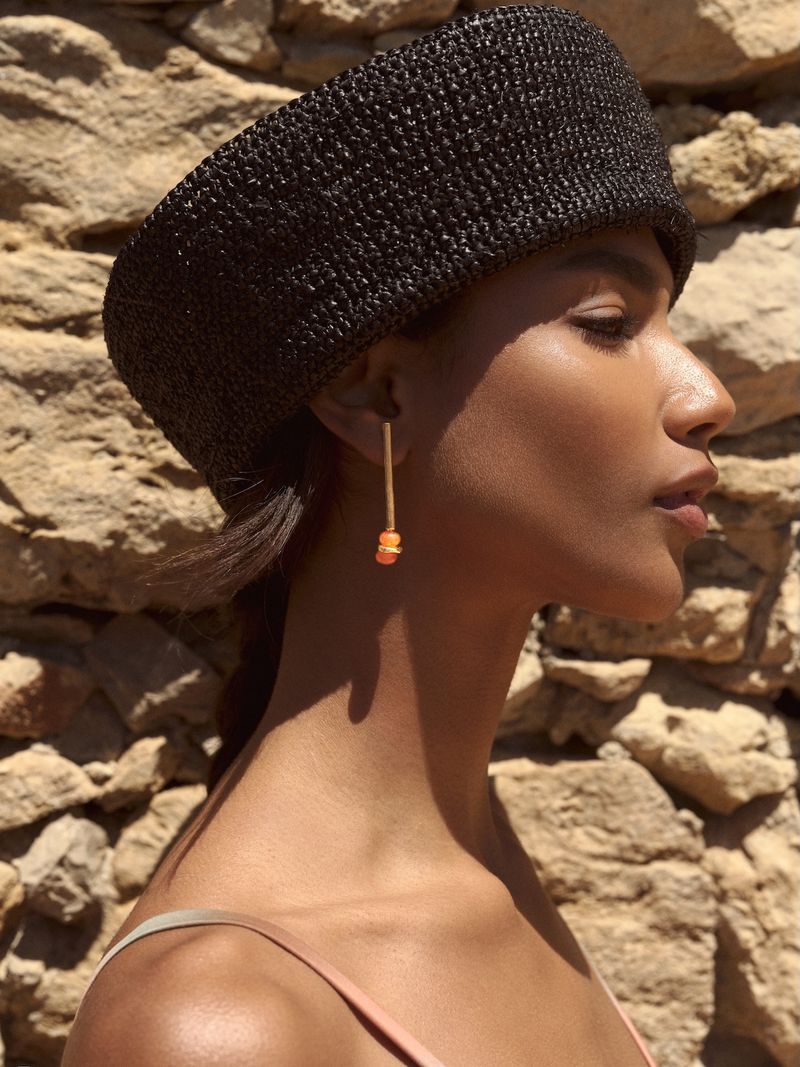 Profile portrait of a model wearing a dark woven hat and drop earrings against a rustic stone wall