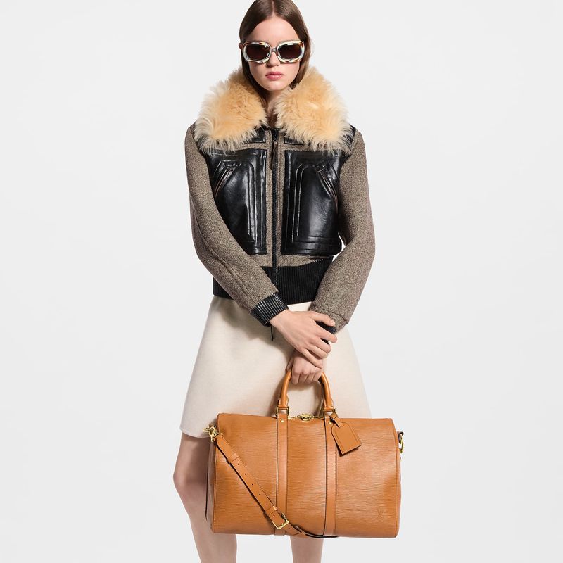 Model wearing sunglasses and a faux-fur collared jacket holding a tan leather duffel bag against a white background