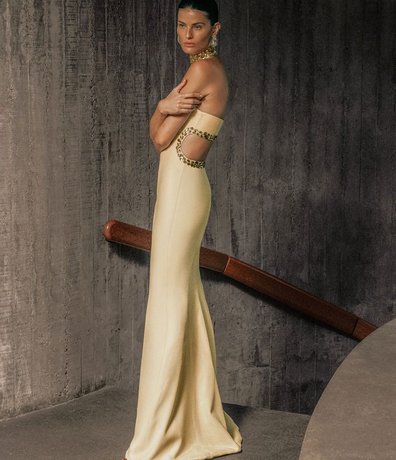Isabeli Fontana in a yellow strapless cutout evening gown standing against a textured concrete wall.