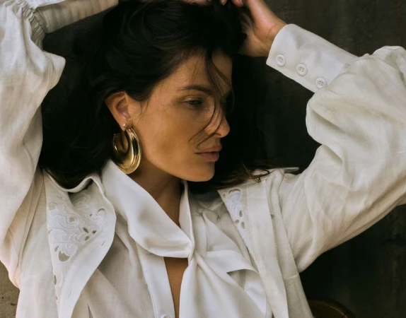 Isabeli Fontana in a white blouse with large gold hoop earrings and her arms raised behind her head for Closet Delux Summer 2027.