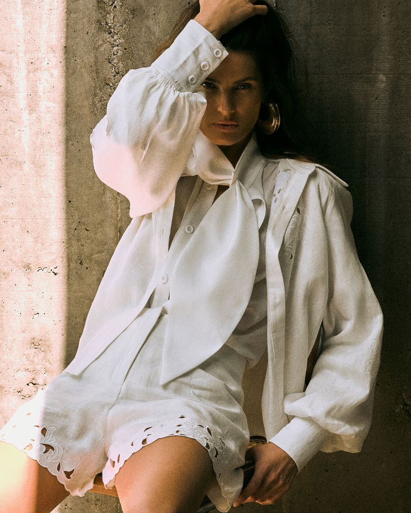 Model Isabeli Fontana wearing a white blouse and shorts against a textured concrete wall.