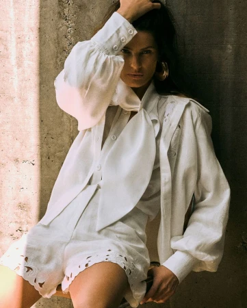 Model Isabeli Fontana wearing a white blouse and shorts against a textured concrete wall.