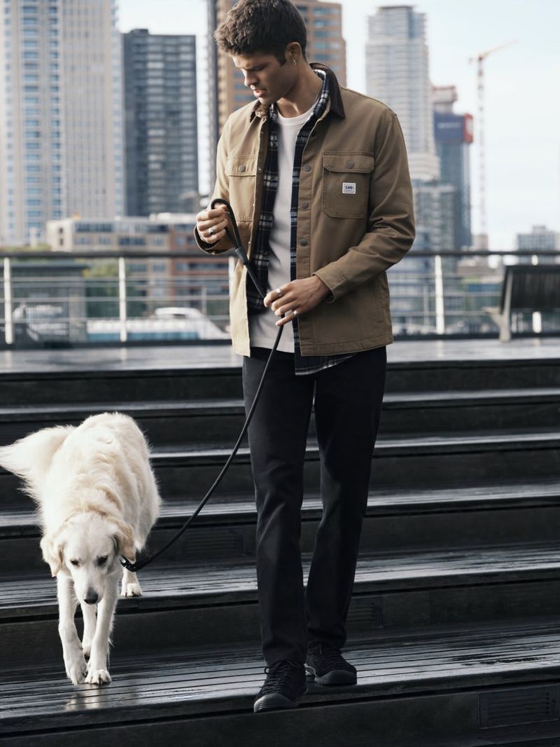 Man walking a cream-colored dog down outdoor steps wearing a brown jacket, plaid shirt, and dark jeans.