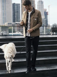 Man walking a cream-colored dog down outdoor steps wearing a brown jacket, plaid shirt, and dark jeans.