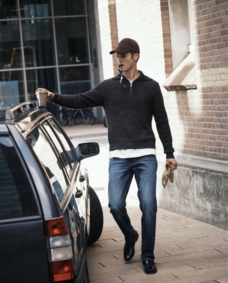 A man wearing blue jeans and a dark zip-up sweater leans against a dark car next to a brick building facade.