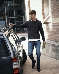 A man wearing blue jeans and a dark zip-up sweater leans against a dark car next to a brick building facade.