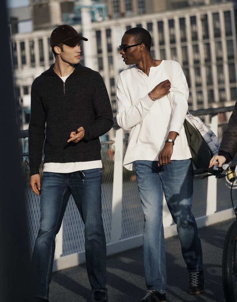 Two men walking and talking outdoors in a city setting wearing denim jeans and casual shirts