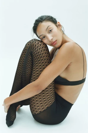 Woman wearing black fishnet tights and black underwear sitting with knees drawn up against a white background.
