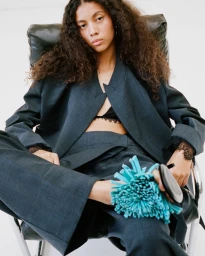 Woman with long curly hair wearing a dark grey suit jacket and holding a blue fringed handbag while seated in a black leather chair.