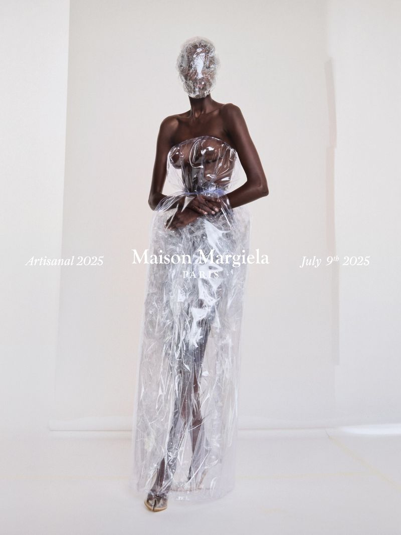 A tall, slender, dark-skinned fashion model presents an avant-garde ensemble featuring a clear plastic dress and head covering against a plain white backdrop.