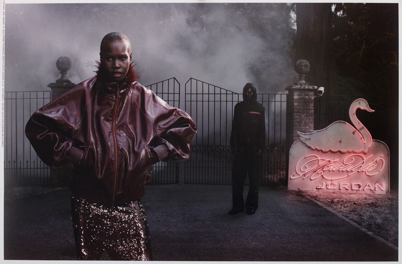 Model in burgundy leather jacket and sequin skirt standing before gates with a neon swan sign for Mowalola x Jordan