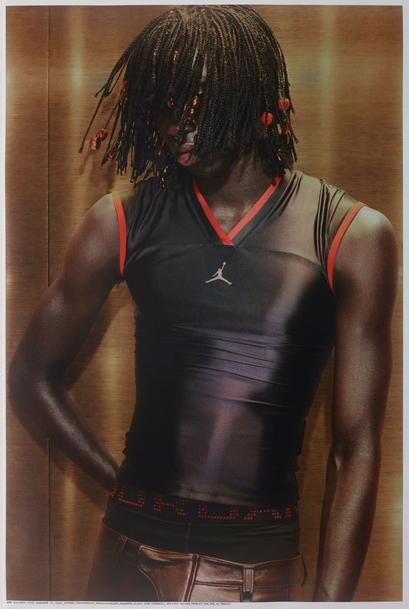 Black model with braided hair wearing a black sleeveless Jordan top with red trim against a warm metallic wood panel background.