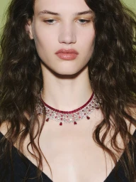 Close-up portrait of a woman with long dark wavy hair wearing an elaborate diamond and ruby necklace against a solid green background