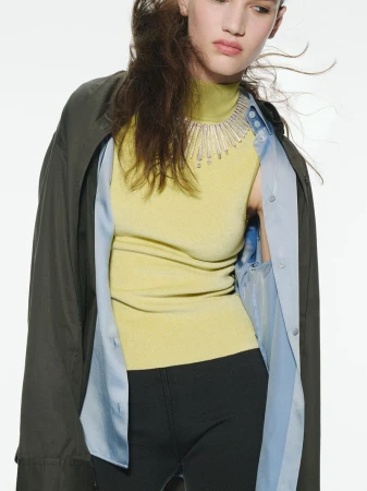 Model wearing a yellow mock-neck top, blue shirt, dark coat, and statement necklace against white