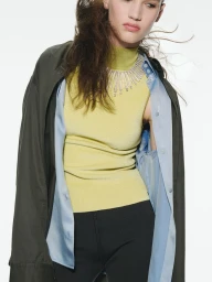 Model wearing a yellow mock-neck top, blue shirt, dark coat, and statement necklace against white