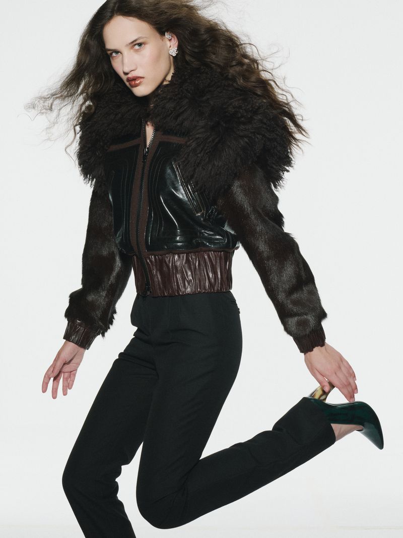 Model with long dark curly hair wearing a dark furry jacket and black trousers against a white seamless background