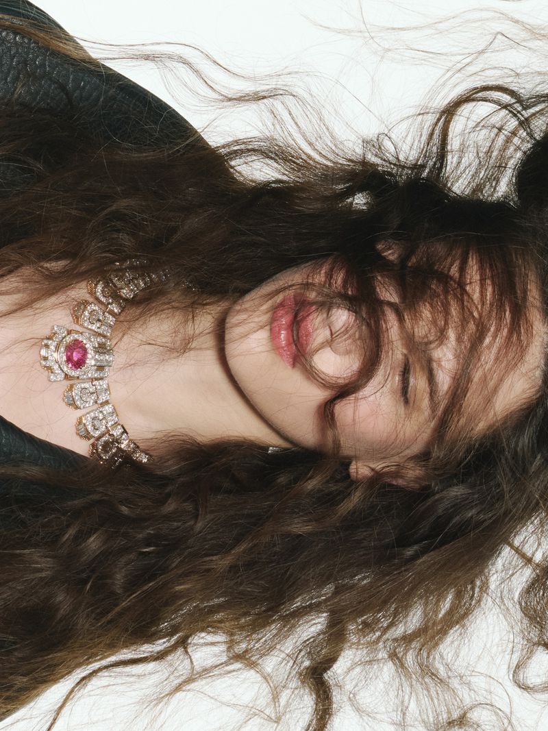 Close-up portrait of a woman wearing an ornate gem-set necklace and dark jacket against a plain white background