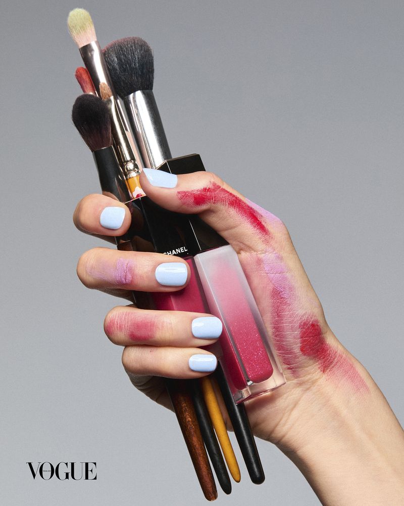 Hand holding makeup brushes and lip gloss with blue manicured nails and cosmetic swatches on skin