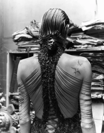 Monochrome rear view of a model with short wet-look hair, wearing a dark laced halter gown with sculpted face ornament and shoulder tattoos.