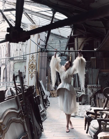Model in white ruffled couture dress holding overhead beams inside a warehouse filled with antique furniture and architectural elements