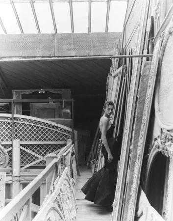 Model wearing a dark evening gown standing among stacked architectural frames and wooden panels.