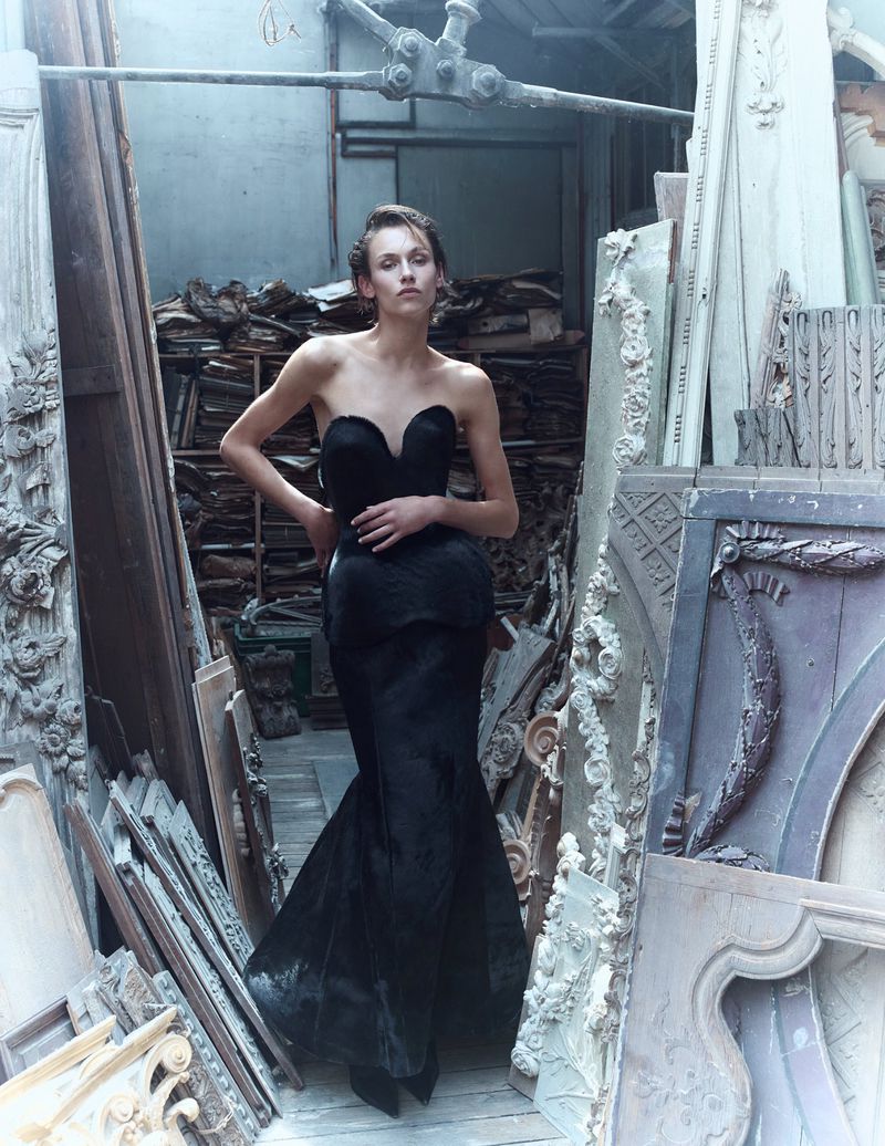 Model wearing a structured black strapless gown standing in an architectural workshop surrounded by carved wooden panels