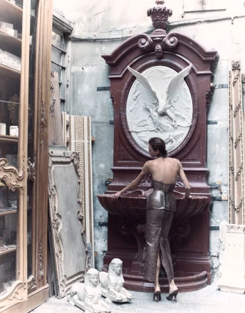 Model wearing structured snakeskin-pattern gown standing with her back to camera in an antique-filled workshop with classical reliefs.