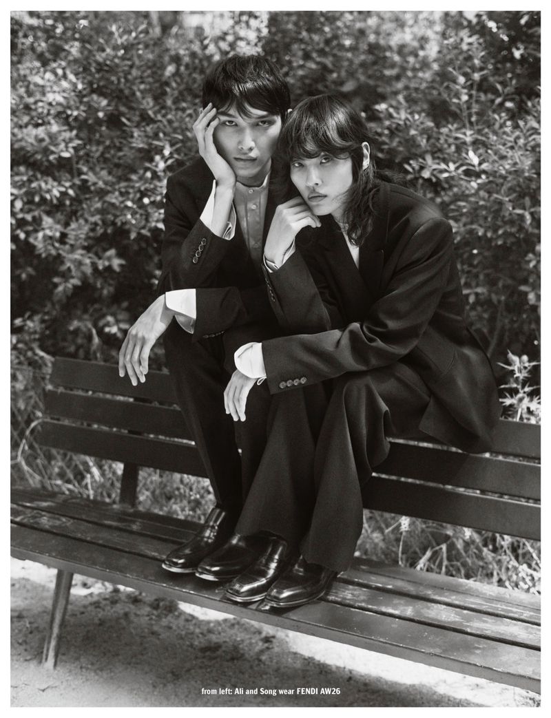 Two models wearing dark black suits seated together on an outdoor wooden park bench amidst lush foliage.