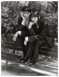 Two models wearing dark black suits seated together on an outdoor wooden park bench amidst lush foliage.