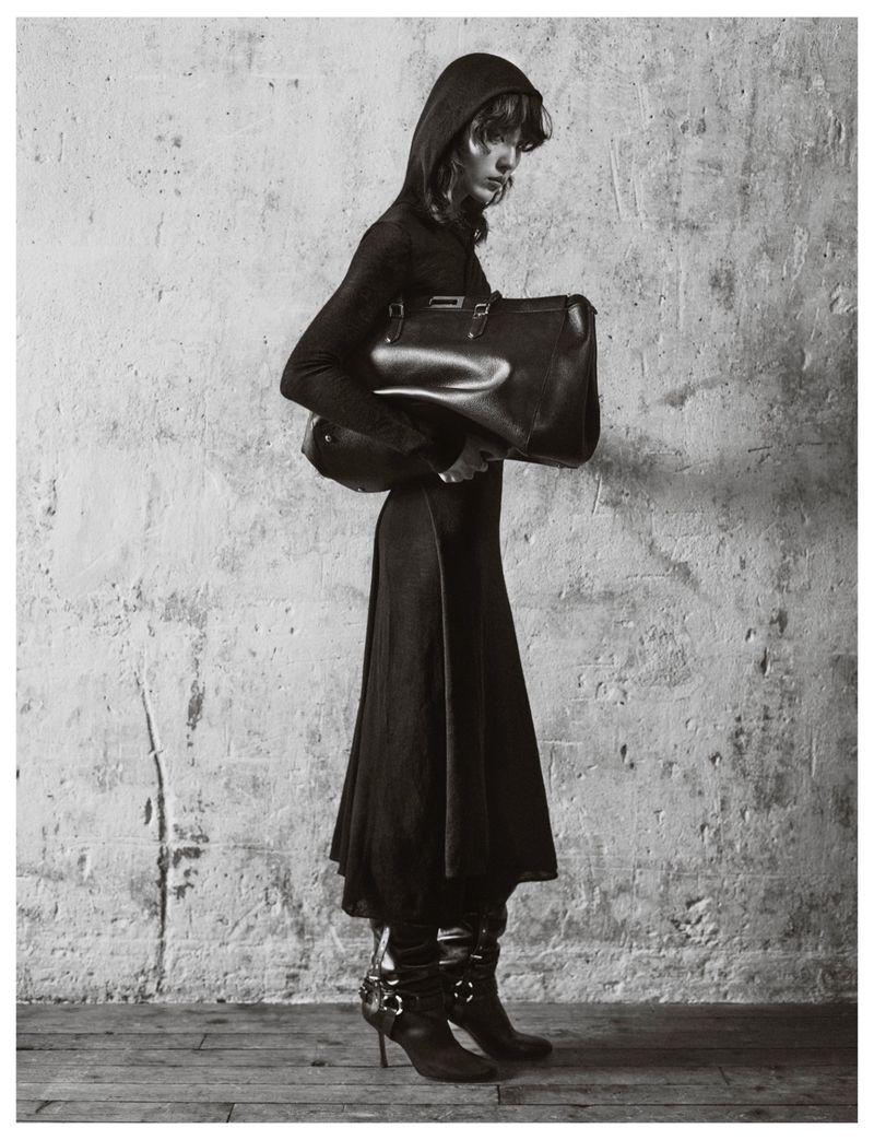 Model in a black hooded dress holding a large leather handbag against a textured concrete wall.