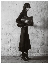 Model in a black hooded dress holding a large leather handbag against a textured concrete wall.