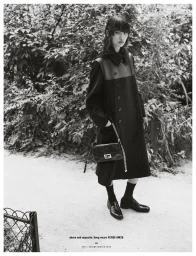 Model Song Ah Woo wearing a long dark coat and holding a shoulder bag on a path bordered by trees.