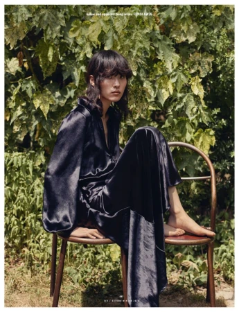 Model Song Ah Woo wearing a long black satin dress, seated on a chair outdoors in front of leafy foliage