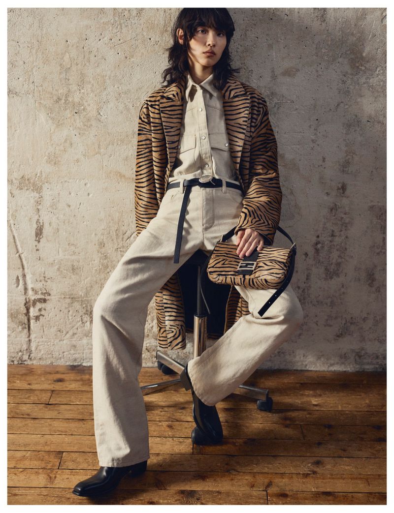 Model seated on a stool wearing a tiger print coat, matching bag, and light pants against a textured concrete wall.