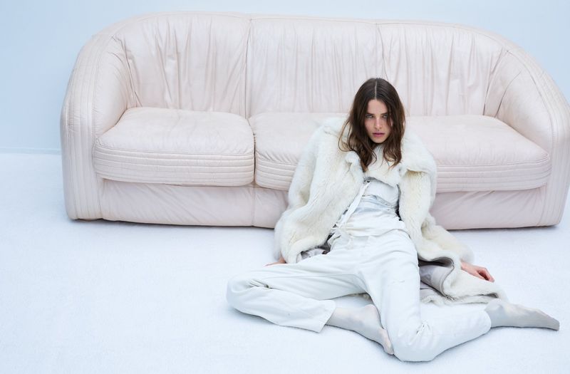 Model seated on the floor wearing a white coat and trousers in front of a white leather sofa.