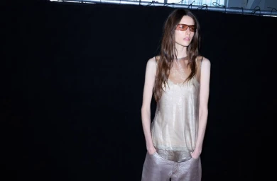 Model wearing tinted sunglasses and a sheer metallic top against a black backdrop