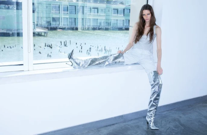 Model wearing metallic silver knee-high boots and grey tank top seated on a windowsill with a waterfront view.