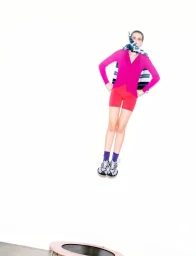 Model in mid-air wearing a vibrant magenta cardigan, red shorts, and a patterned scarf against a plain white studio background.