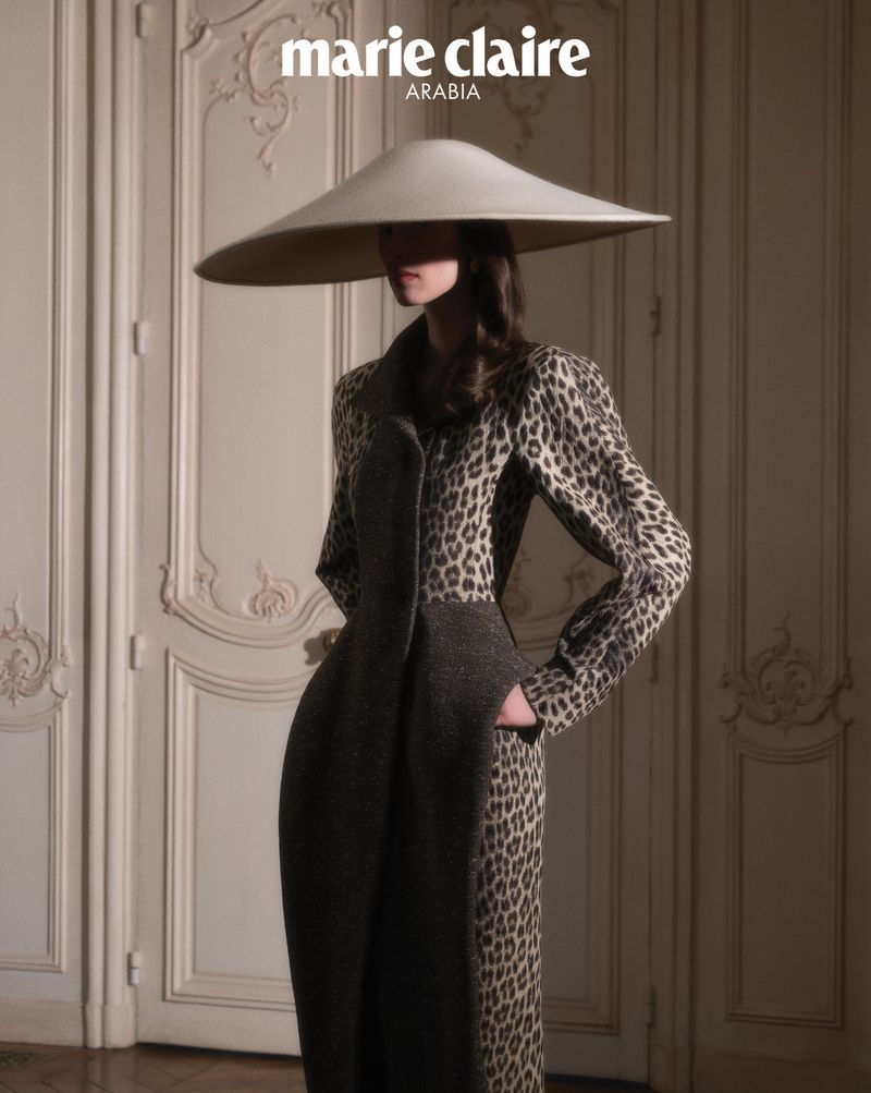 Model wearing a wide-brimmed cream hat and patterned coat standing in front of paneled double doors