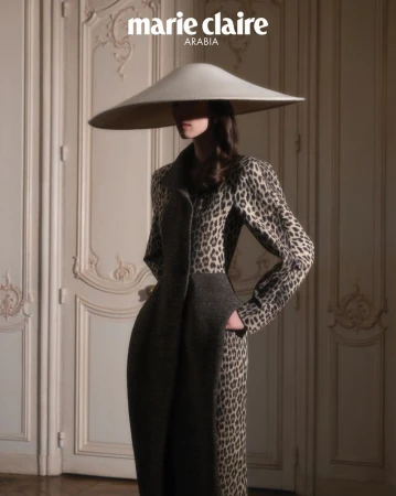 Model wearing a wide-brimmed cream hat and patterned coat standing in front of paneled double doors