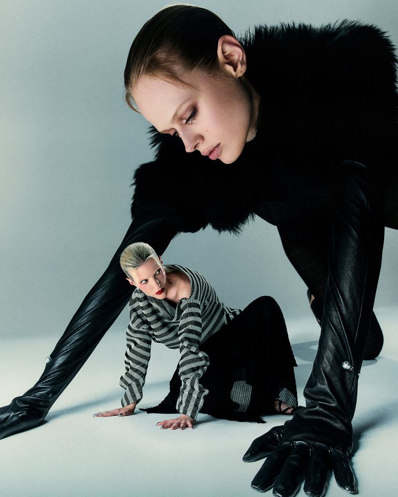 Two women pose on a white studio floor. One is kneeling in a striped sweater, the other is behind, in black fur and black gloves.