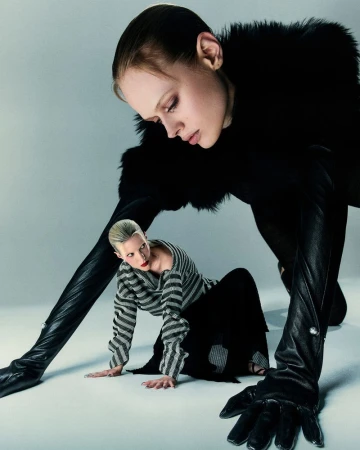 Two women pose on a white studio floor. One is kneeling in a striped sweater, the other is behind, in black fur and black gloves.