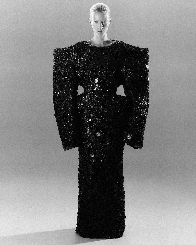 A fair-skinned model poses in a full-length shot, wearing a dark, textured, dress-like garment with circular button accents in a studio setting.
